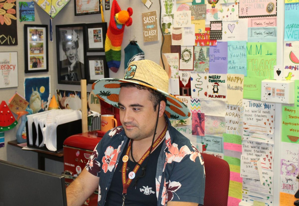 Cameron Martin, English 10 Honors and English 11 College Prep teacher, works at his desk in a beach-themed spirit day outfit. Cards from students line the wall behind him.
