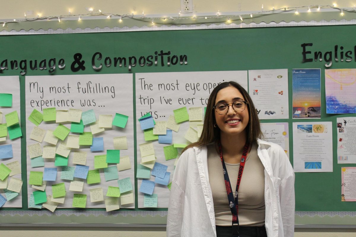 Anna Regula, English 9 Honors and AP Language and Composition teacher, stands in front of a wall of student work with string lights twinkling overhead. Regula joined the school's English department this year. 