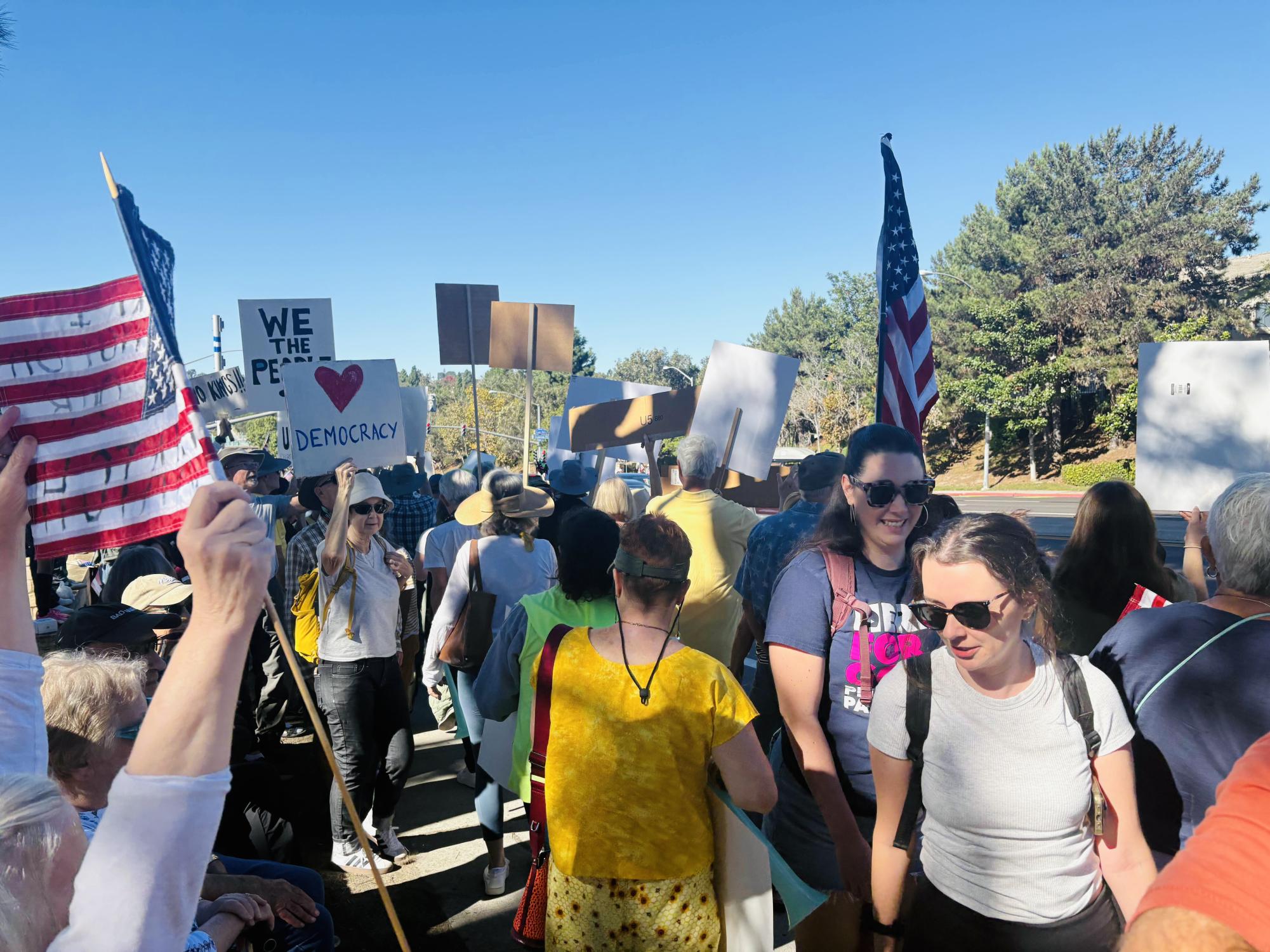Community members in front of Del Mar Highlands Town Center sporting yellow attire weave up and down the sidewalk on Del Mar Heights Road. Protesters held up American flags and signs with slogans such as “We the People” and “No Kings.”